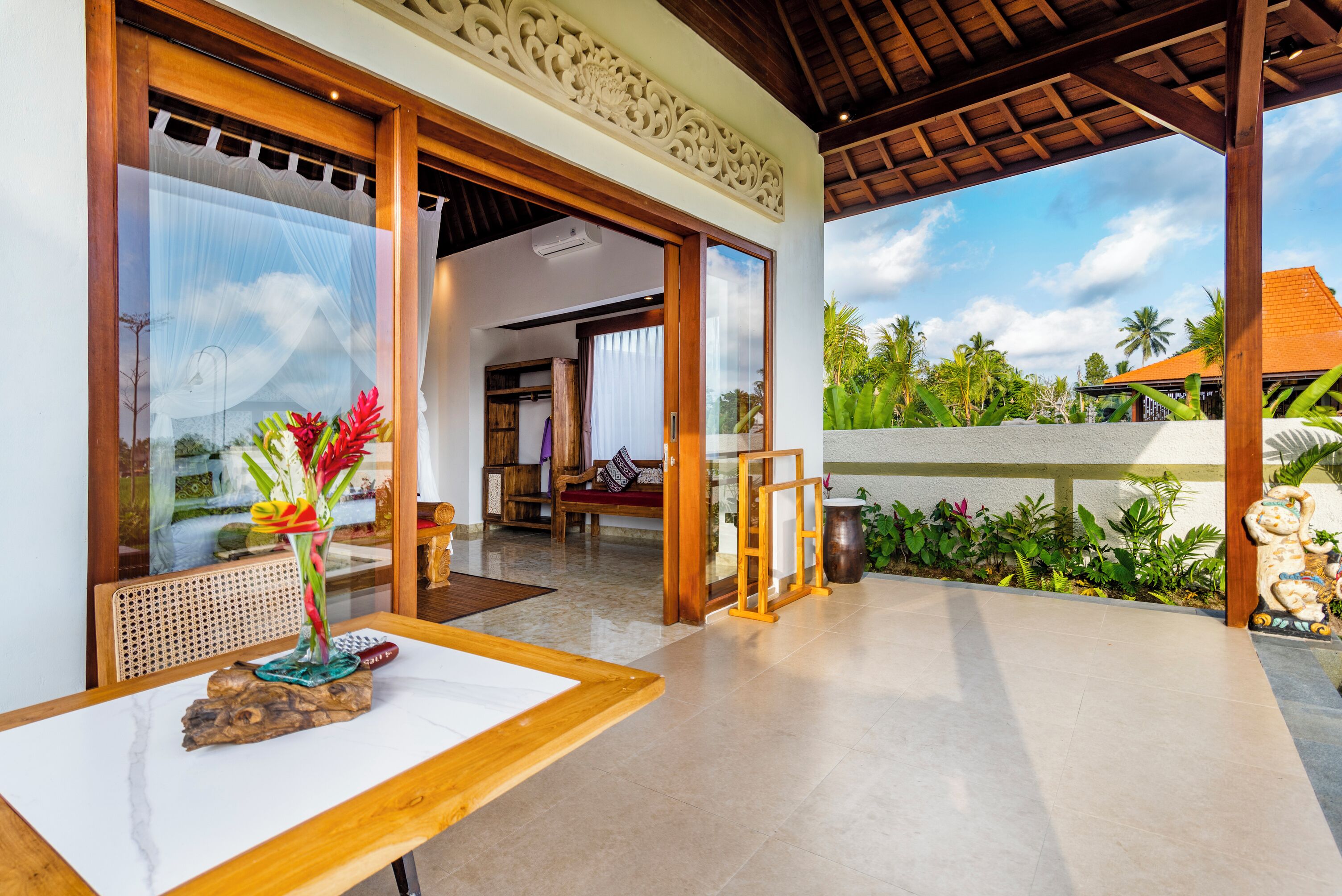 Un vaste espace extérieur agrémenté d’une table en bois, d’un vase décoratif et offrant une vue sur un paysage tropical, avec des palmiers et un bâtiment en arrière-plan.