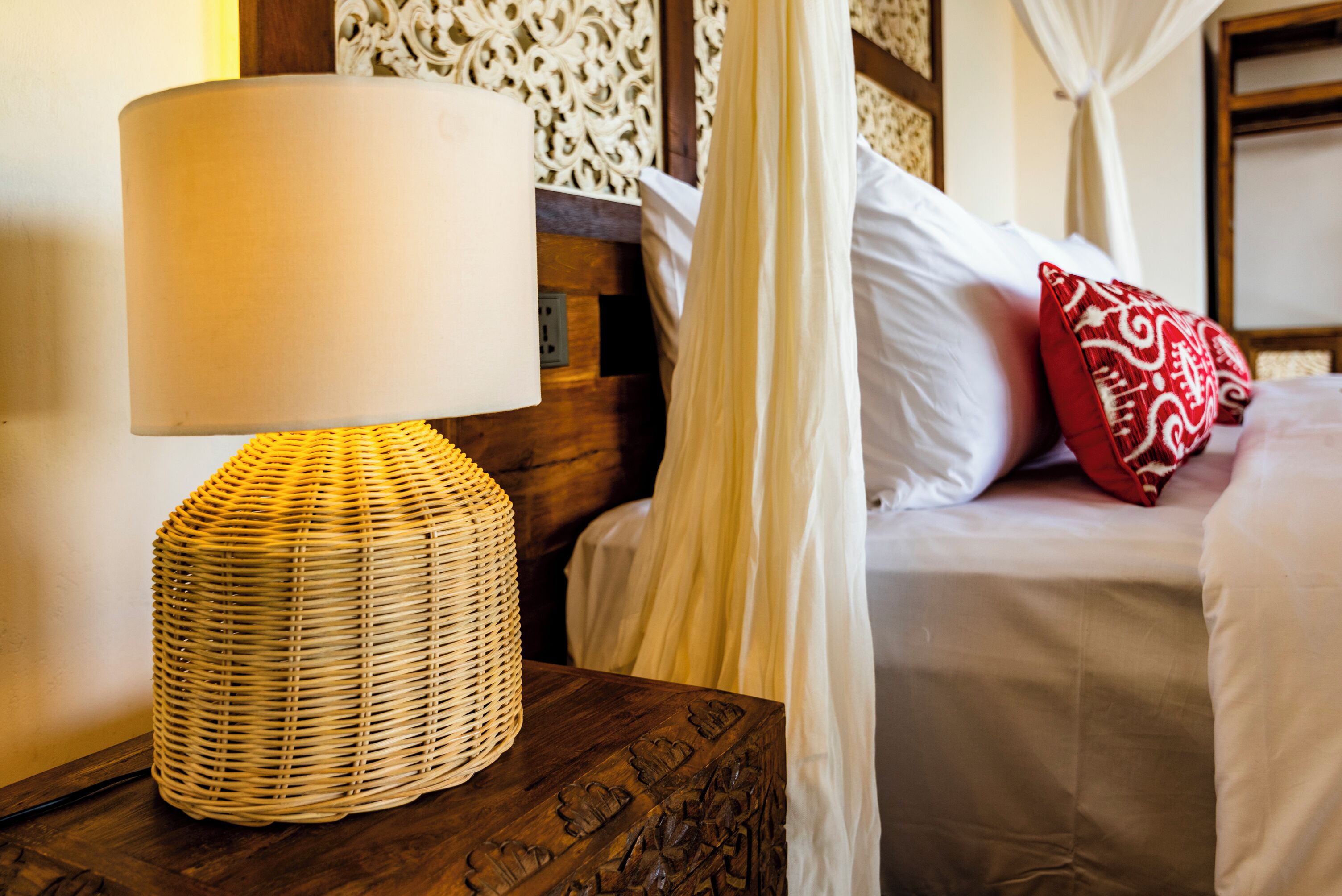 A neatly made bed with white linens and a red patterned pillow, a woven lamp on a wooden nightstand, and a canopy bed with white curtains.