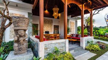 Un espacio de estar al aire libre techado, con esculturas de piedra, una pérgola de madera y abundante vegetación.