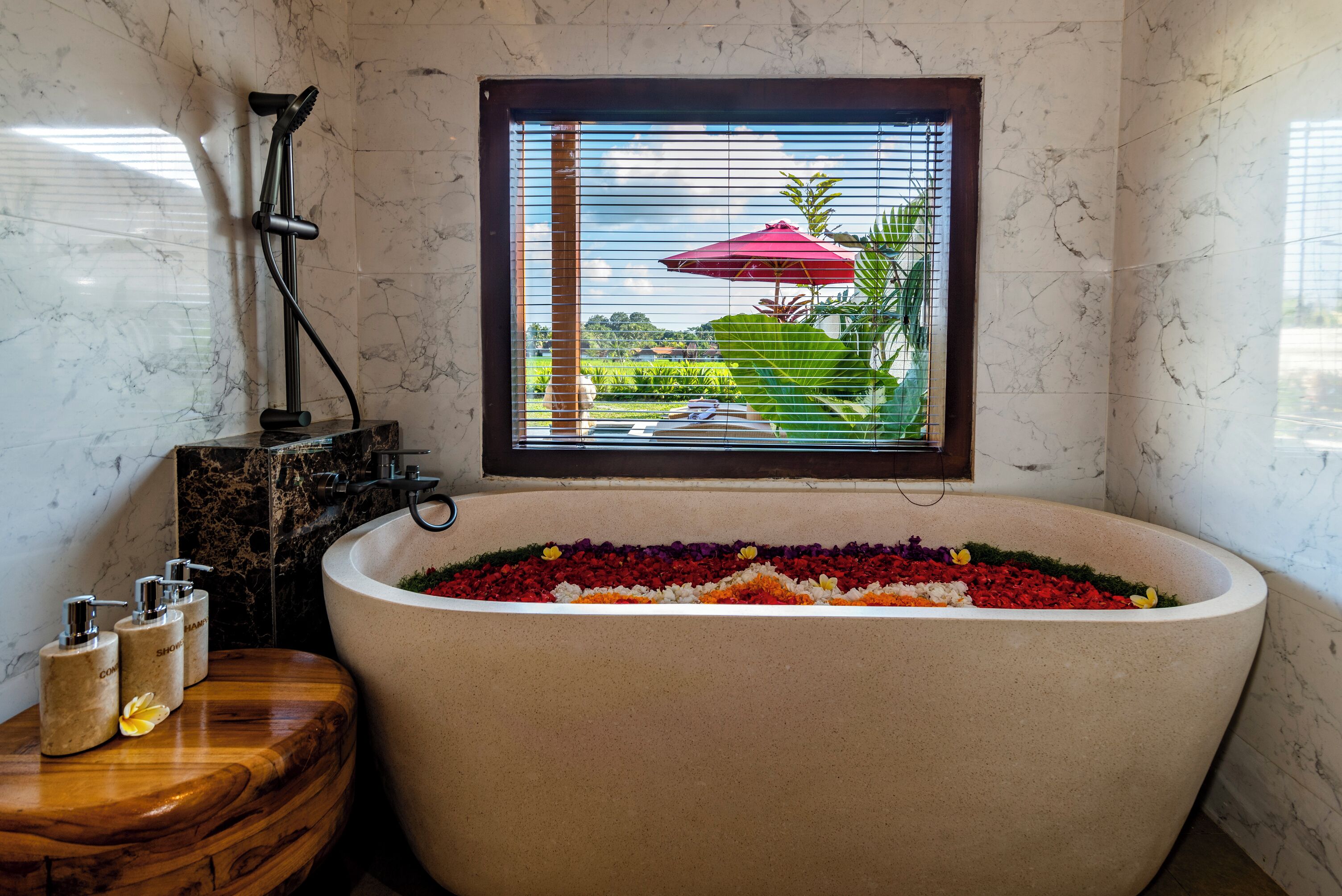 A bathroom with a large bathtub filled with colorful flowers, a window with a view of a garden, and a wooden side table with toiletries.