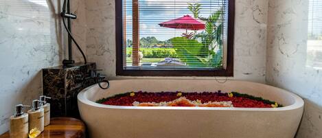 A bathroom with a large bathtub filled with colorful flowers, a window with a view of a garden, and a wooden side table with toiletries.