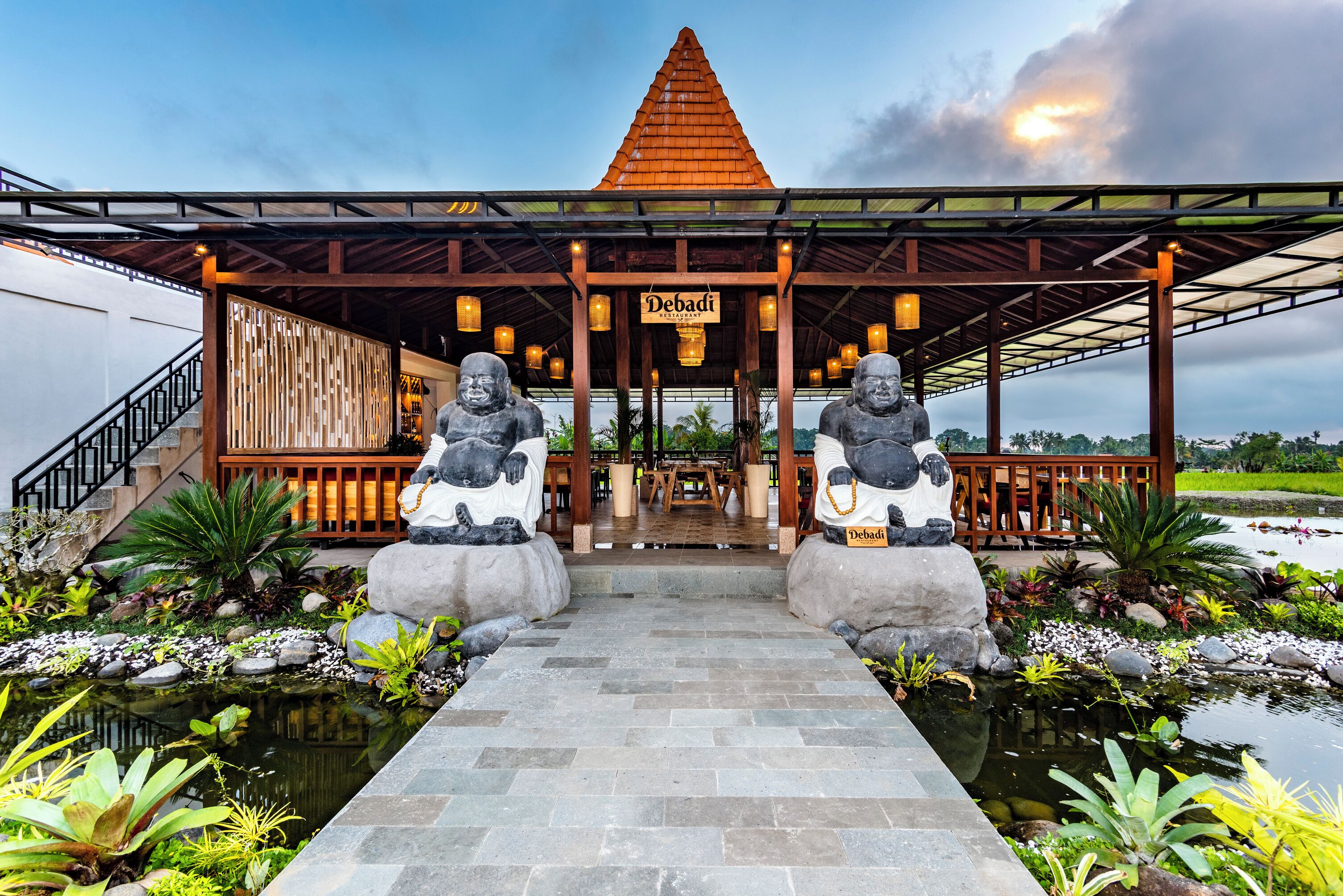 A restaurant with a covered entrance, two stone statues, and a pond with tropical plants.
