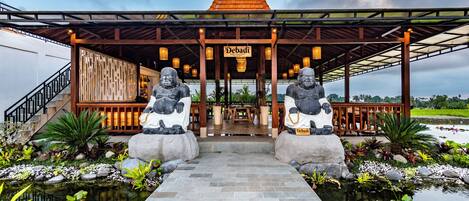 A restaurant with a covered entrance, two stone statues, and a pond with tropical plants.