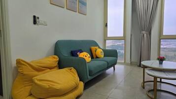 A living room with a sofa, yellow cushions, a round table with a flower, and a view through large windows.