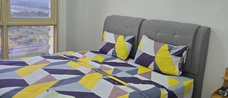 Un lit double avec une couvre-lit à motifs géométriques, une tête de lit rembourrée de couleur grise et une vue sur le paysage urbain, visible à travers une grande fenêtre.