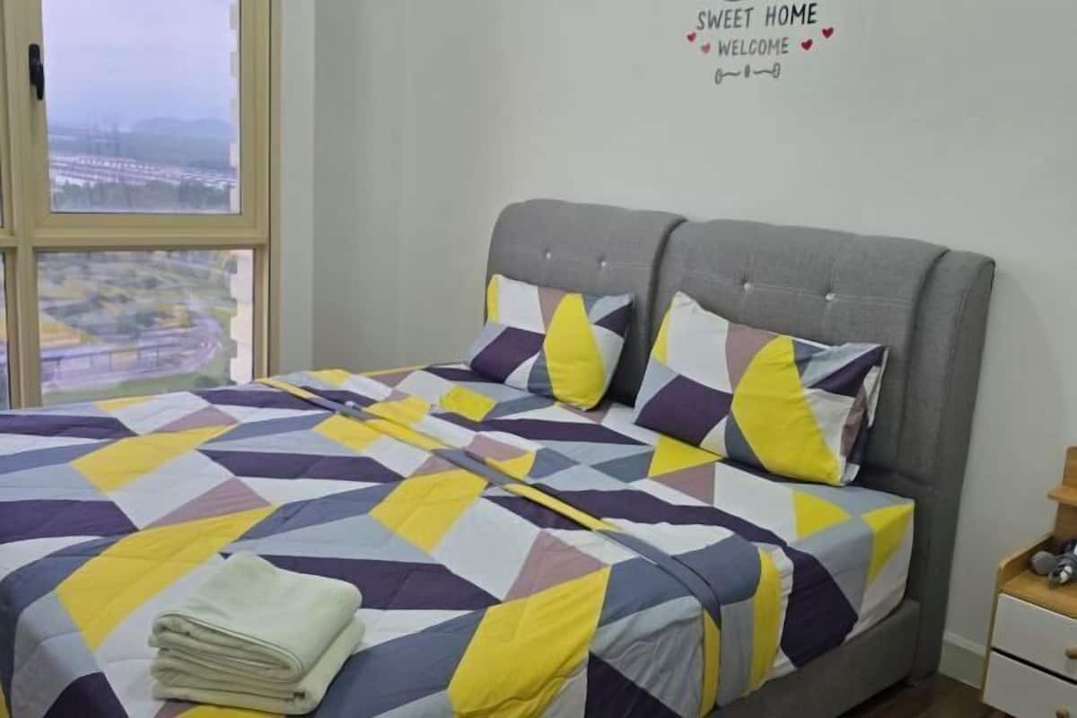 A bed with a geometric patterned bedspread, a grey upholstered headboard, and a view of a cityscape through a large window.