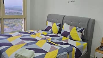 A bed with a geometric patterned bedspread, a grey upholstered headboard, and a view of a cityscape through a large window.