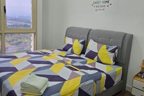 Een bed met een bedspreider met geometrisch patroon, een grijs gestoffeerd hoofdbord en een uitzicht op een stadsgezicht door een groot raam.