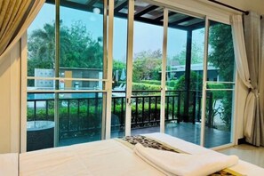 Une chambre dotée d’une grande porte-fenêtre coulissante donnant sur un balcon avec vue sur la verdure et une piscine.