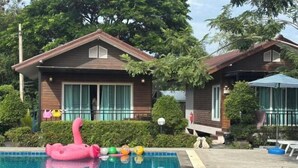 Une piscine agrémentée d’un flotteur en forme de flamant rose et de ballons colorés, entourée de deux maisons en bois, de haies vertes et d’arbres.