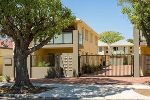 Un edificio residencial de dos plantas con un camino de entrada cerrado por una reja, un árbol en la parte delantera y un cielo despejado.