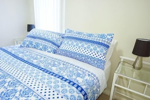 Uma cama com colcha estampada, uma mesa de cabeceira com abajur e uma janela com cortinas.