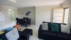 A living room with a black sofa, a dining table with chairs, a kitchen area, and a door with a window. - Unit Close to Airport, Hospital, Shops & Transport (Midland)