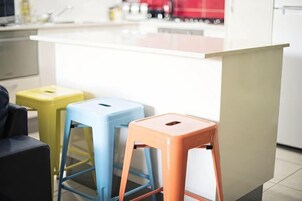 Una cocina con encimera blanca, tres taburetes de colores y un armario color arena.