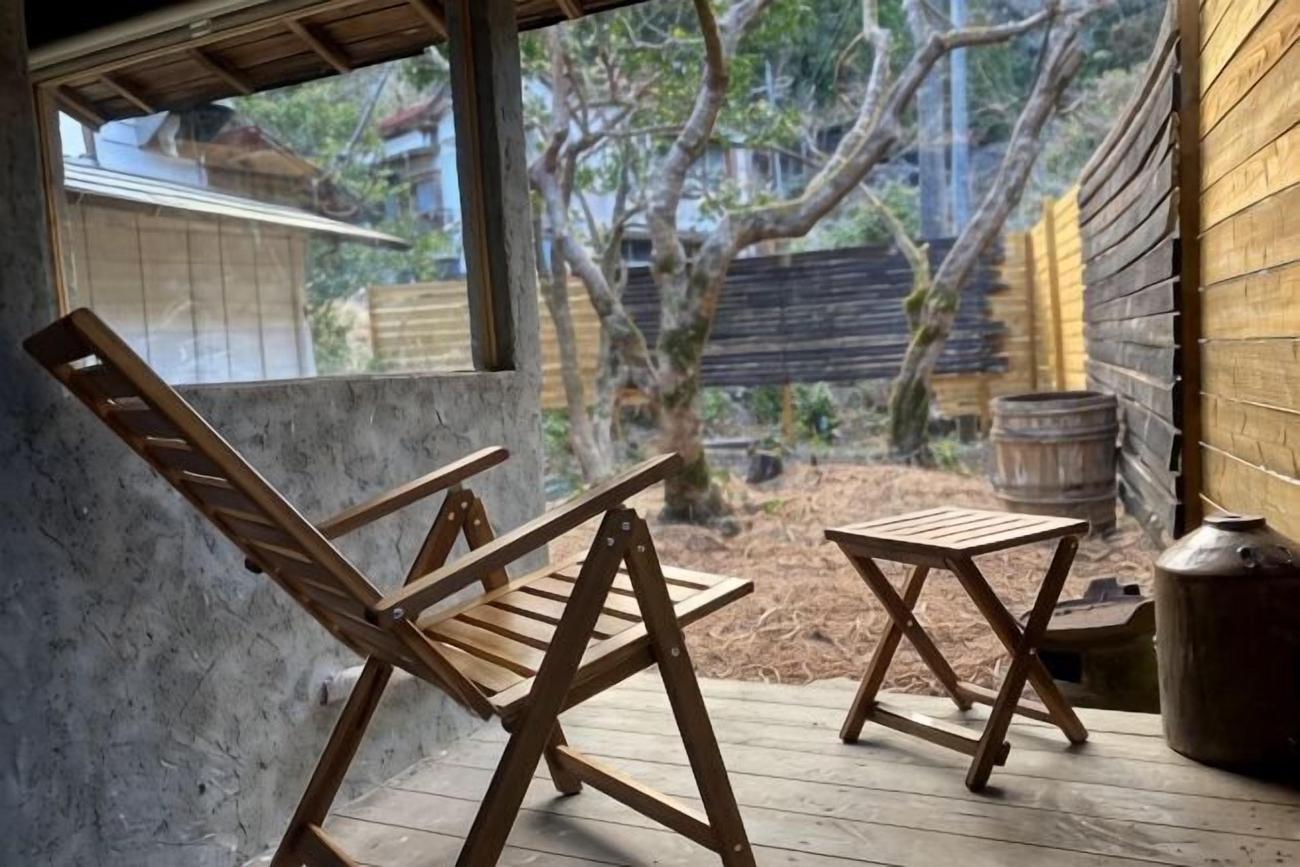 Une terrasse en bois avec deux chaises et une petite table, une fenêtre et une vue sur un arbre et une clôture.
