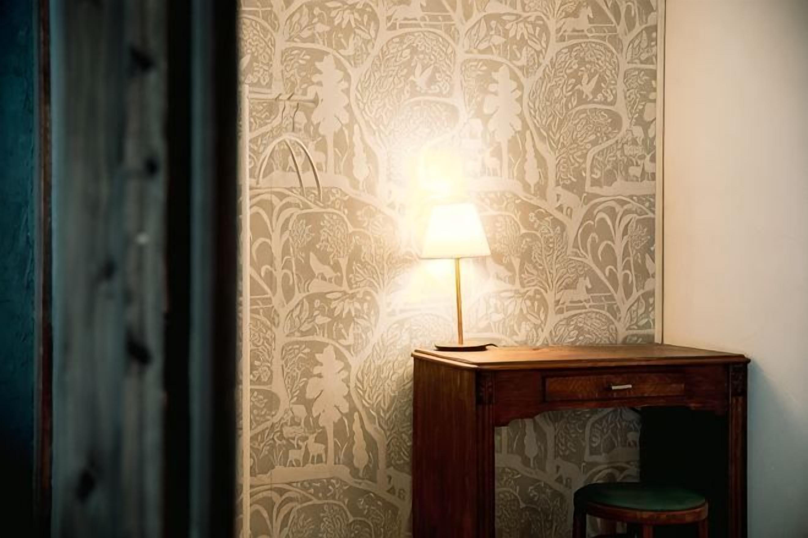 A room with a patterned wallpaper, a wooden desk with a lamp, and a chair.