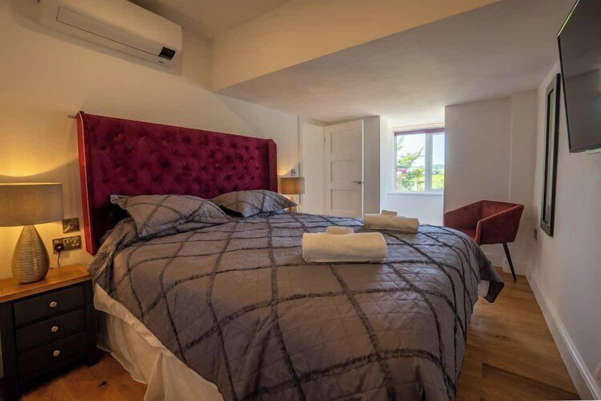 A bedroom with a large bed, a nightstand, a red chair, and a window with a view of greenery.