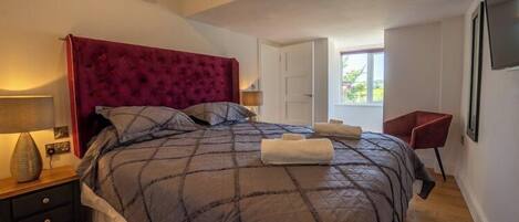 Ein Schlafzimmer mit einem großen Bett, einem Nachttisch, einem roten Sessel und einem Fenster mit Blick auf Grünflächen.