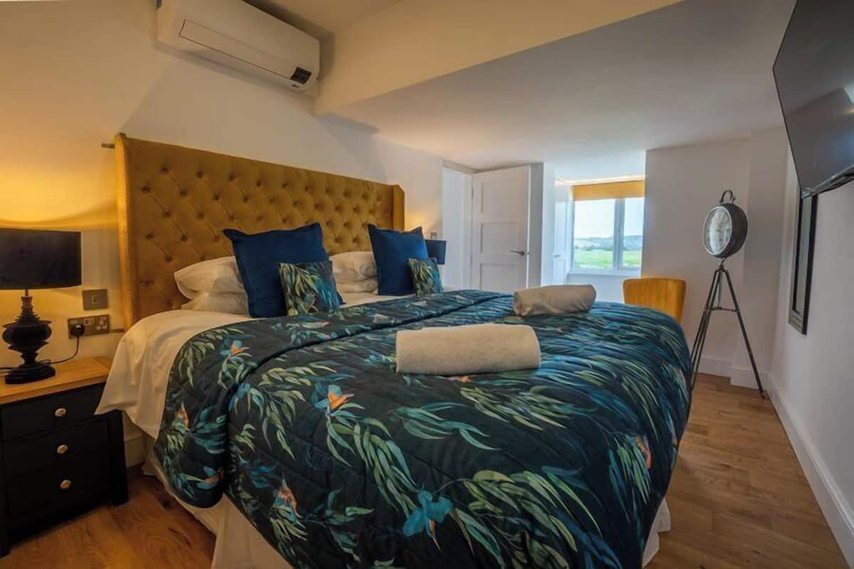 A bedroom with a large bed, a nightstand, a lamp, an air conditioner, a clock, and a window with a view.