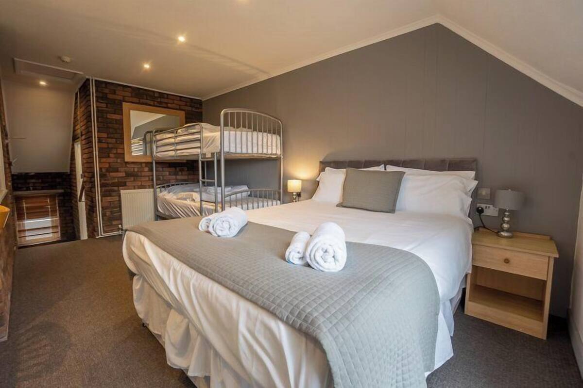 A bedroom with a bunk bed, a large bed with a quilt, a bedside table with a lamp, and a brick accent wall.