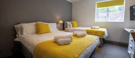 A hotel room with two beds, each with white linens and yellow accents, a bedside table with a lamp, and a window with a view of the outside.