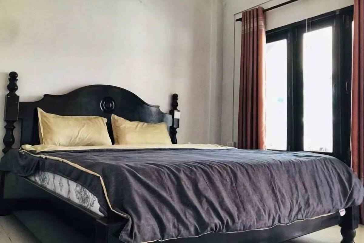 Un lit avec une tête de lit sombre, deux oreillers et une couvre-lit sombre, dans une pièce dotée d’une fenêtre et de rideaux.
