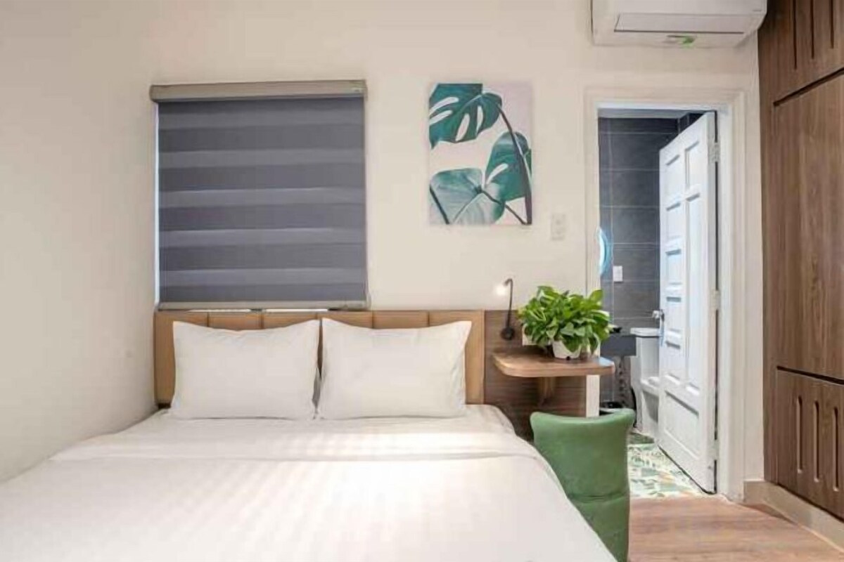 A bedroom with a bed, a bedside table with a plant, a window with blinds, and a door leading to another room.