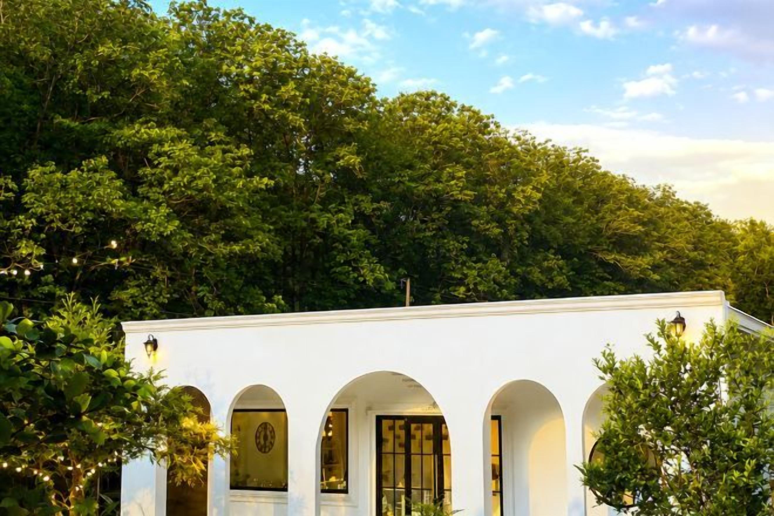 A white building with arched windows and a balcony, surrounded by dense green trees.