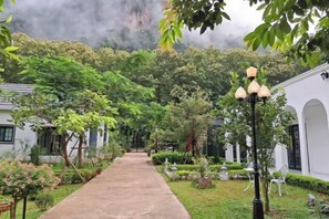 A pathway leading to a building with a traditional architectural style, surrounded by lush greenery and a clear sky. - Vrapat (Chiang Dao)