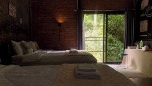 A hotel room with two beds, a large window with a view of greenery, a brick wall, and a small table with a chair. - Vrapat (Chiang Dao)