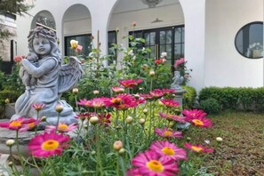 A statue of an angel holding a flower in a garden with pink flowers and a building in the background. - Vrapat (Chiang Dao)
