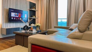 Een moderne woonkamer met een leren bank, een bijzettafeltje met boeken en een vaas, een flatscreen televisie bevestigd aan een houten muur, en een groot raam met gordijnen.