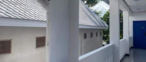 Un pasillo con una pared exterior blanca, una puerta azul y una ventana que da a un edificio con tejado de tejas.