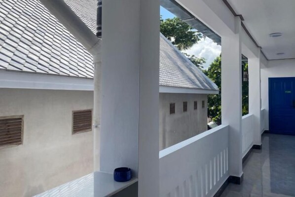A corridor with a white exterior wall, a blue door, and a window overlooking a building with a tiled roof.