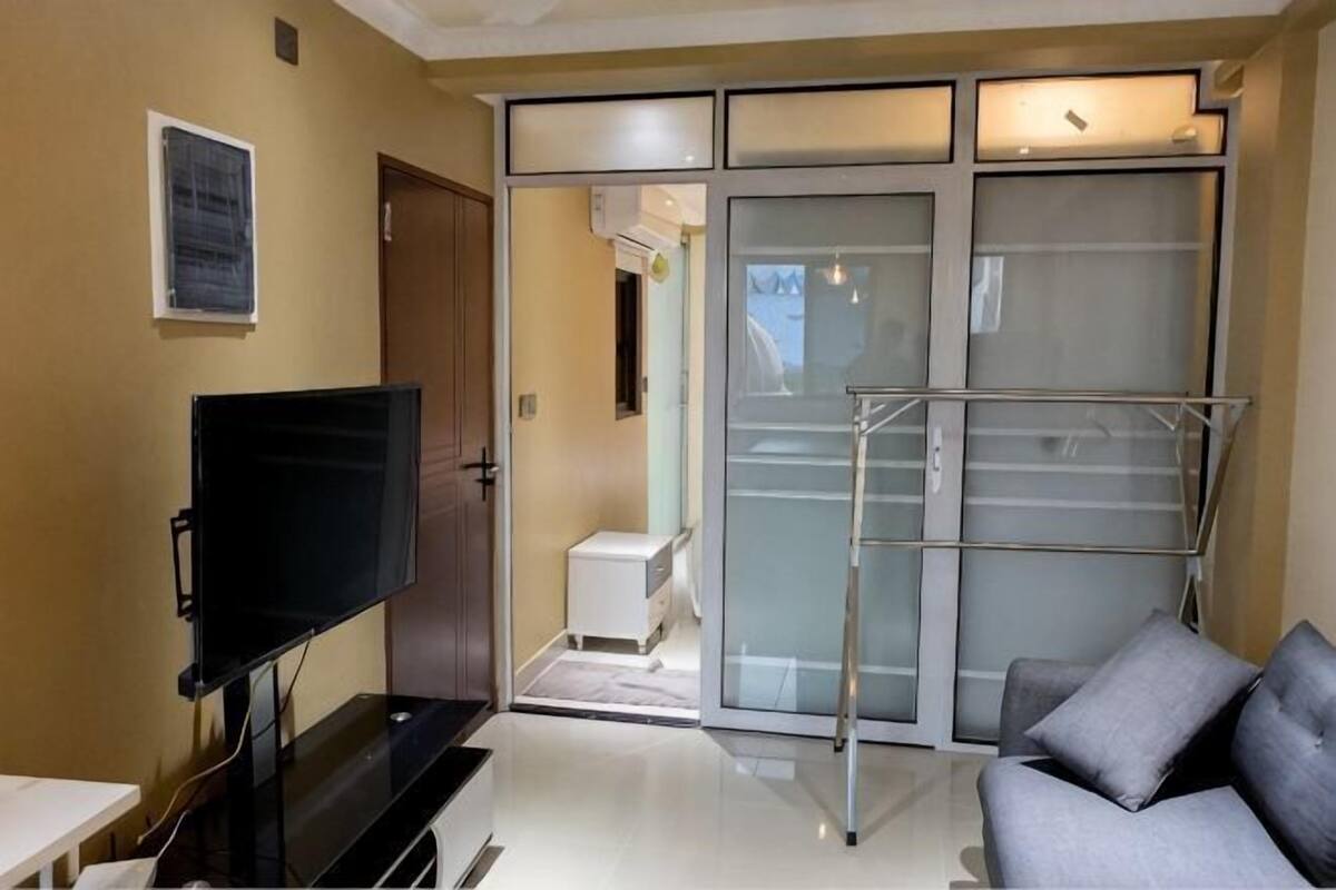 A hotel room with a flat-screen TV, a sliding door closet, a white bedside table, and a grey sofa.