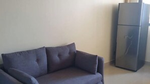 A grey sofa in a room with a large, dark-colored refrigerator against a plain wall. - Fanfini Residence Male’ City (Malé)