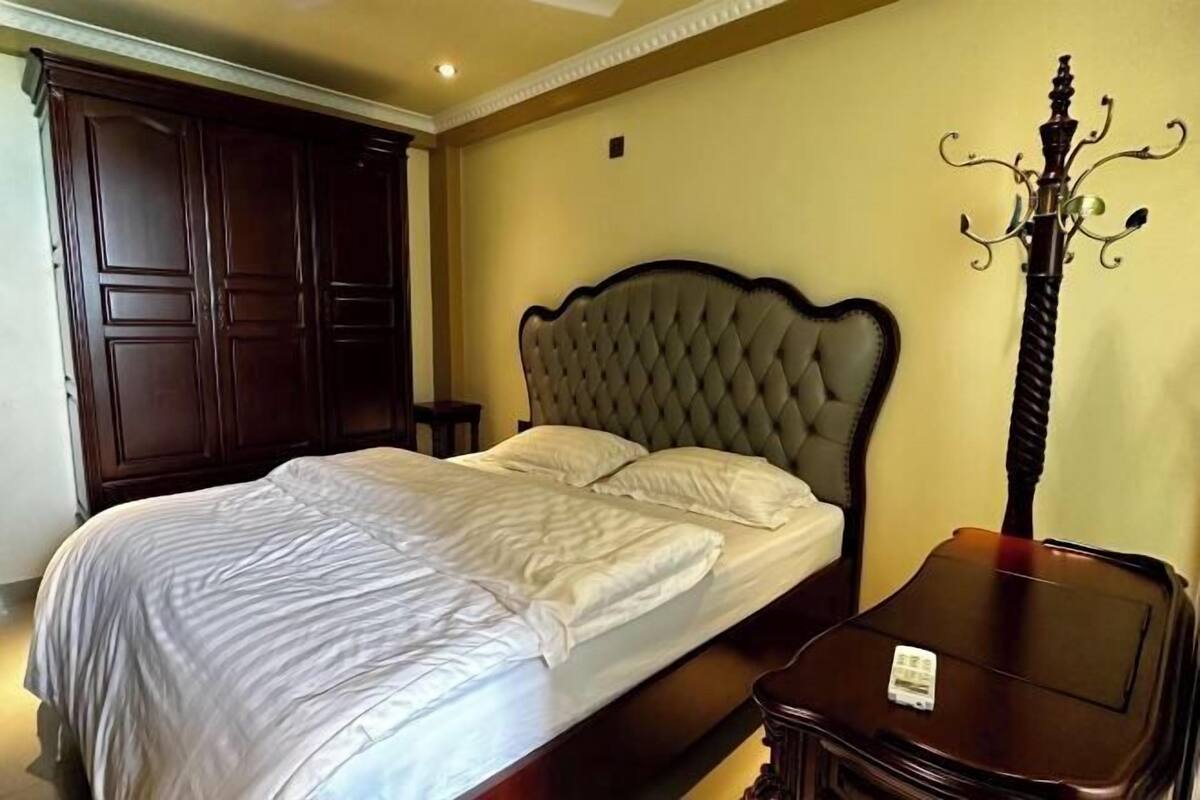 A neatly made bed with a tufted headboard, a dark wooden wardrobe, and a small bedside table with a lamp.