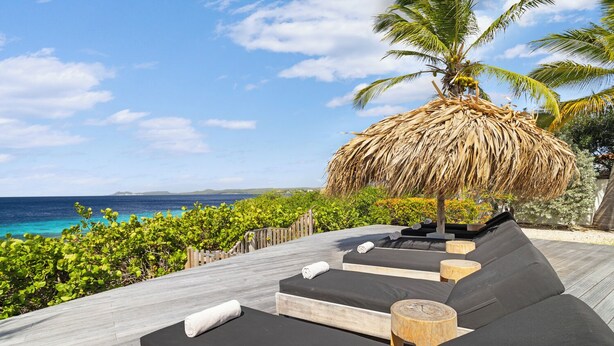 A wooden deck with black cushioned seating, a thatched umbrella, and a view of the ocean. - Stunning Oceanfront Vacation Villa with Private Pool and Beach Access (Kralendijk)