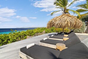 A wooden deck with black cushioned seating, a thatched umbrella, and a view of the ocean. - Stunning Oceanfront Vacation Villa with Private Pool and Beach Access (Kralendijk)