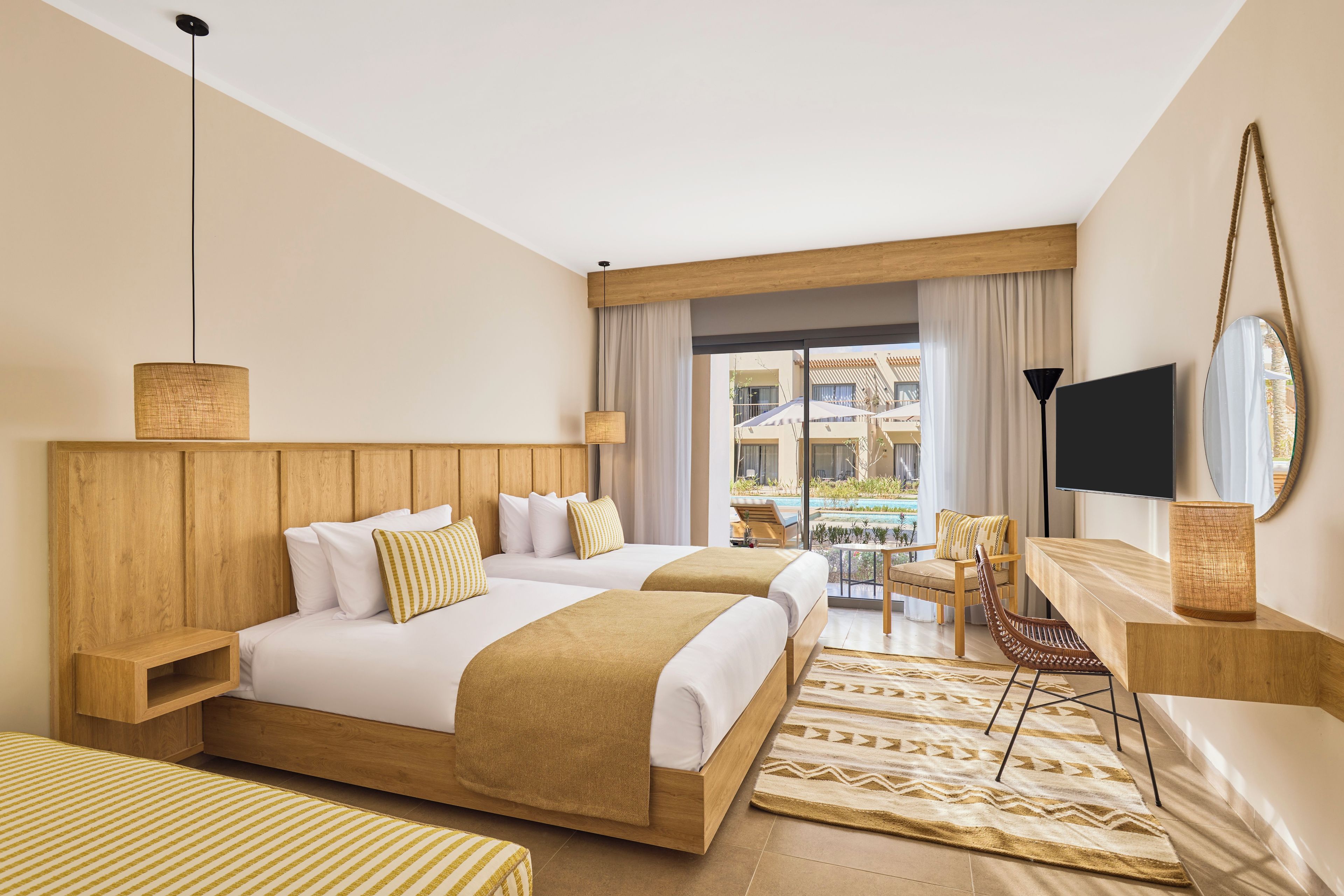 A hotel room with two beds, a TV, a desk, and a view of a pool and buildings.