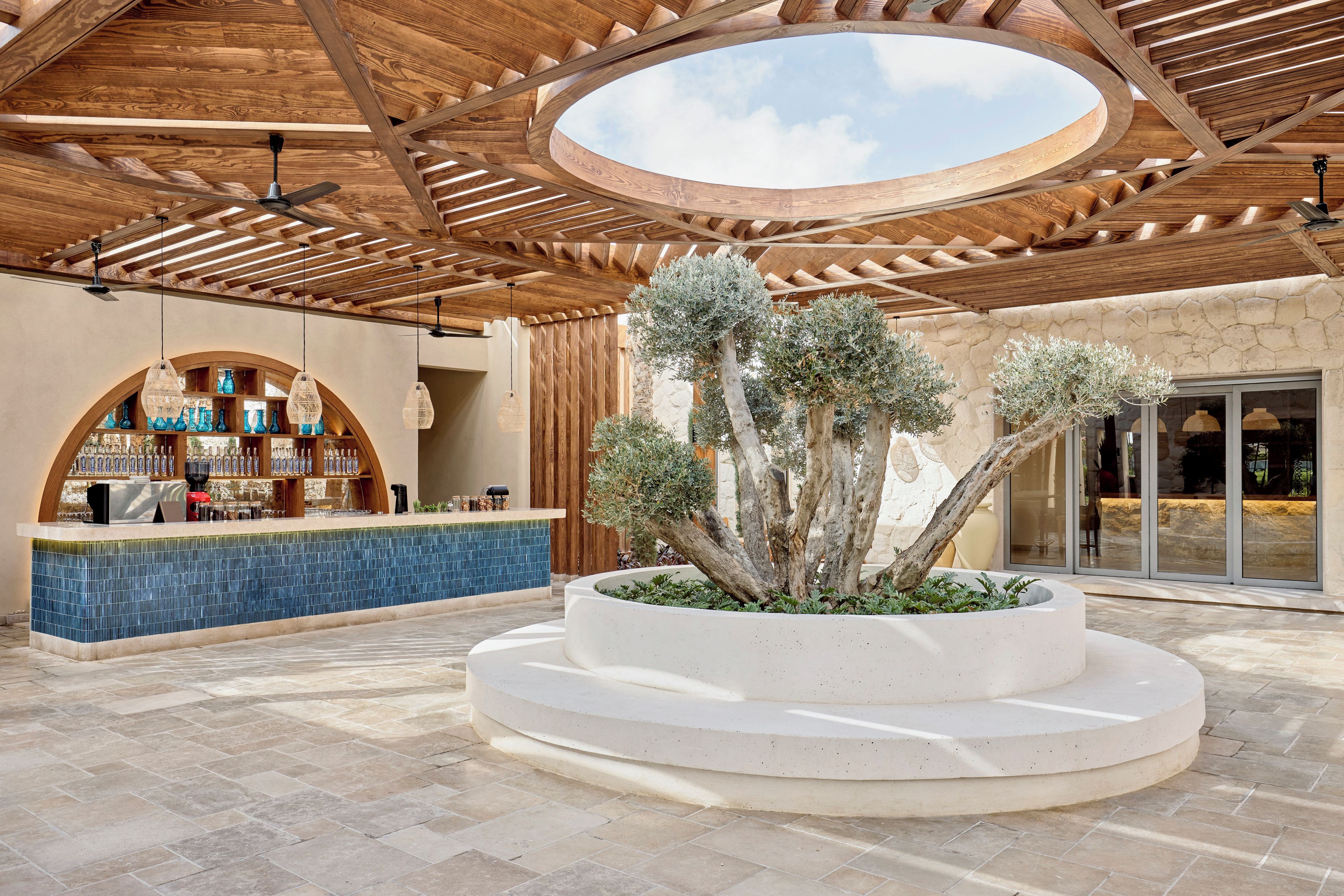 A covered outdoor area with a central planter with trees, a bar area with blue-tiled counter, and a wooden ceiling structure.