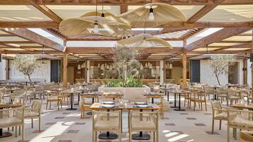 A spacious indoor dining area with wooden tables and chairs, a central plant arrangement, and a high wooden ceiling with natural light.