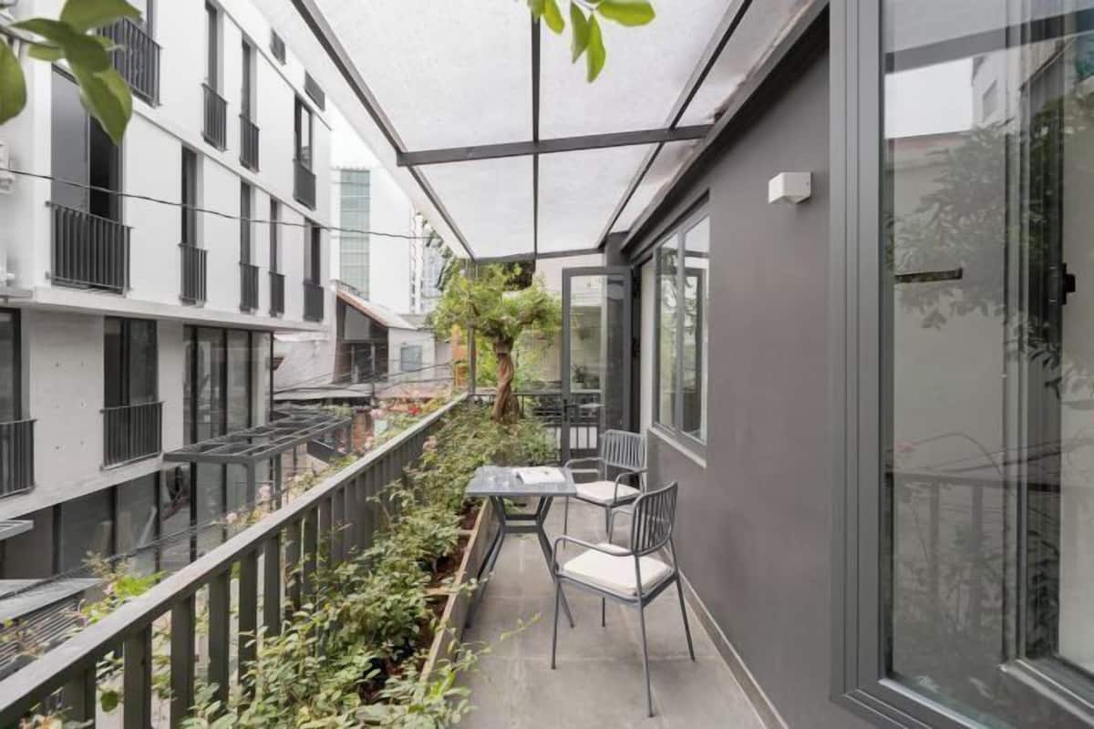 A narrow outdoor corridor with a glass railing, a table with chairs, and a view of adjacent buildings.