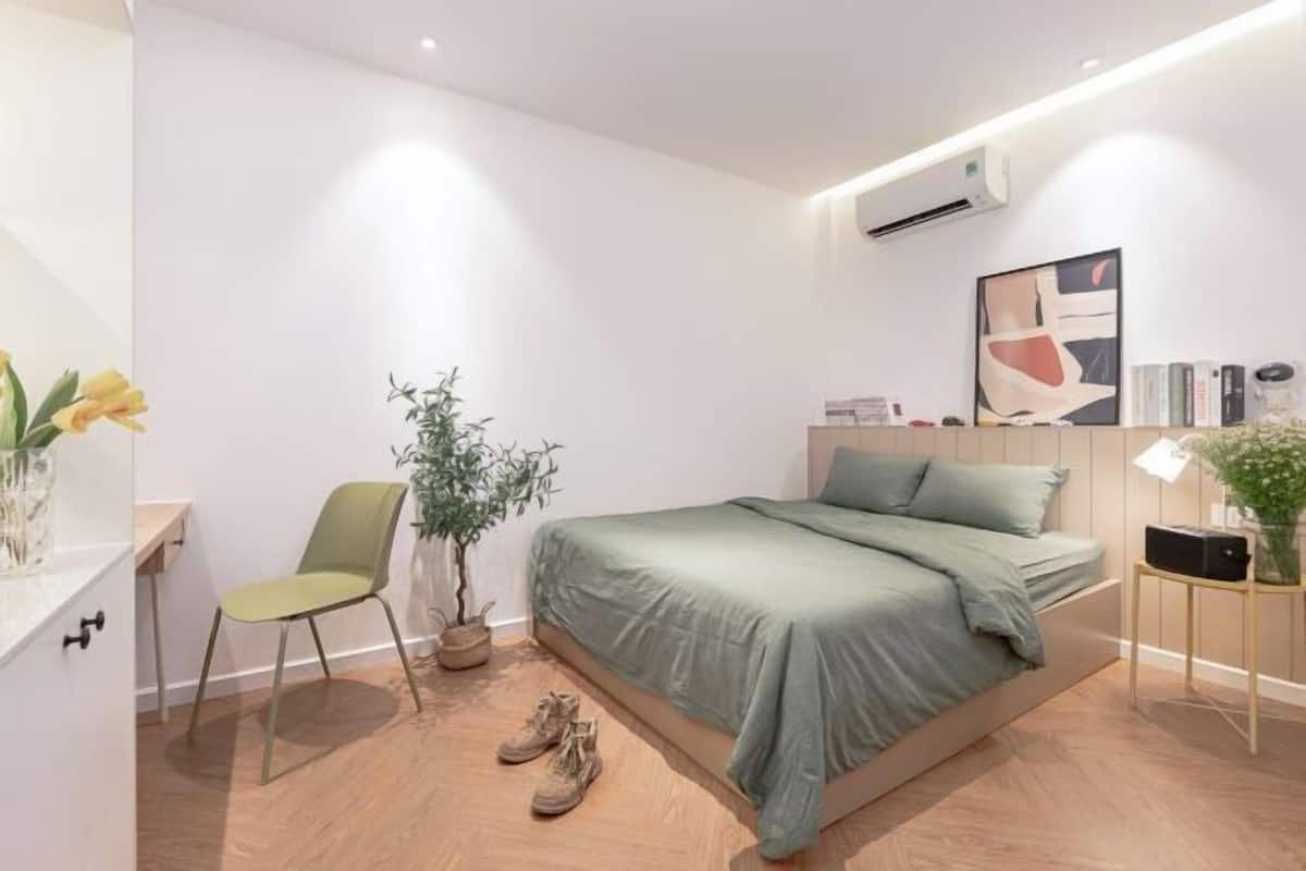 A bedroom with a bed, a chair, a potted plant, a pair of shoes, and a bedside table with a lamp.