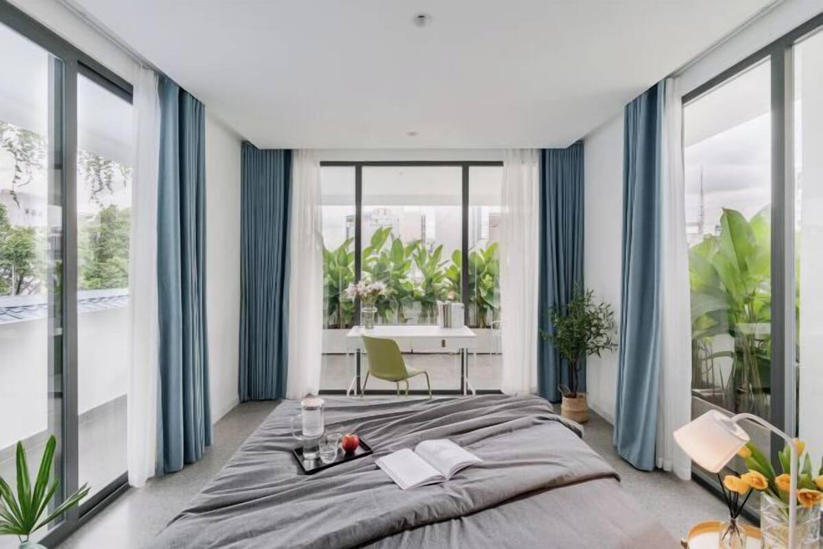 A modern bedroom with a large bed, a dining table, and a view of greenery through large windows.