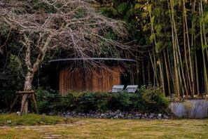 A rustic wooden structure with a curved roof, surrounded by a garden with a tree and a stone wall. - RICKA KATSUURA (Katsuura)