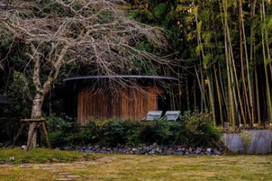 Una estructura rústica de madera con techo curvo, rodeada por un jardín con un árbol y un muro de piedra.