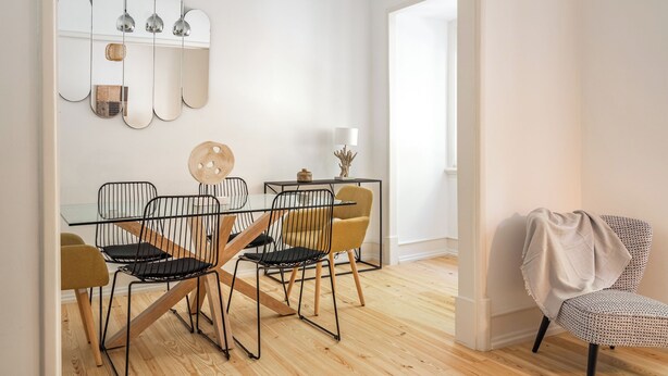 Un coin repas avec une table en verre, des chaises en fil de fer noir et une chaise jaune. Des miroirs sont fixés au mur et des lampes suspendues sont placées au-dessus de la table.