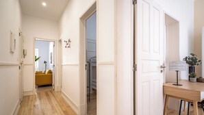 Un couloir doté d’un plancher en bois, de murs blancs et d’une porte donnant sur une pièce où se trouvent un lit et une chaise jaune.