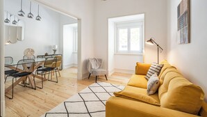 Un salon moderne avec un canapé jaune, un coin repas doté d’une table et de chaises en verre, et une grande fenêtre laissant entrer la lumière naturelle.